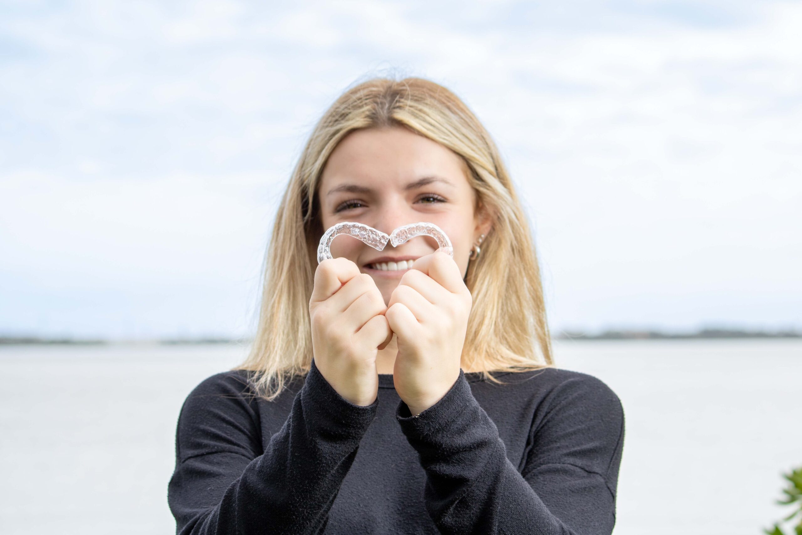 Teenager holding her Invisalign® in Melbourne, FL in the shape of a heart by the water.